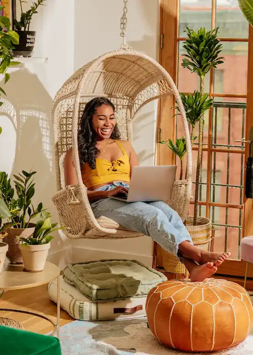 Women smiling and sitting in a wicker swing while typing on her laptop.