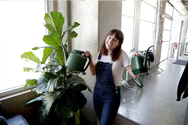 A woman smiles while holding a watering can and waters a plant in the office