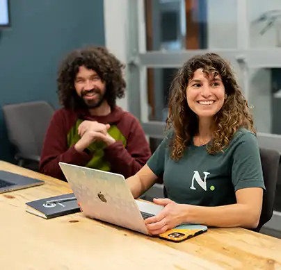 A man and a woman sit behind a laptop at a conference table looking off into the distance and smiling