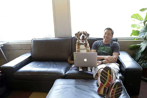 A man sits on the couch next to a medium size dog while holding a laptop in front of the dog like the dog is writing code