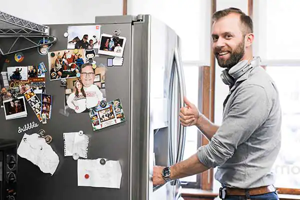 A man with a beard smiles while standing in the office kitchen next to the refrigerator