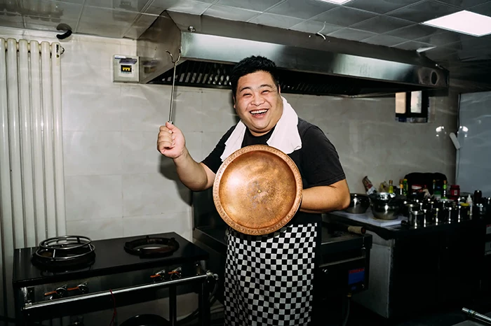 Chef smiling holding copper lid and kitchen utensil.