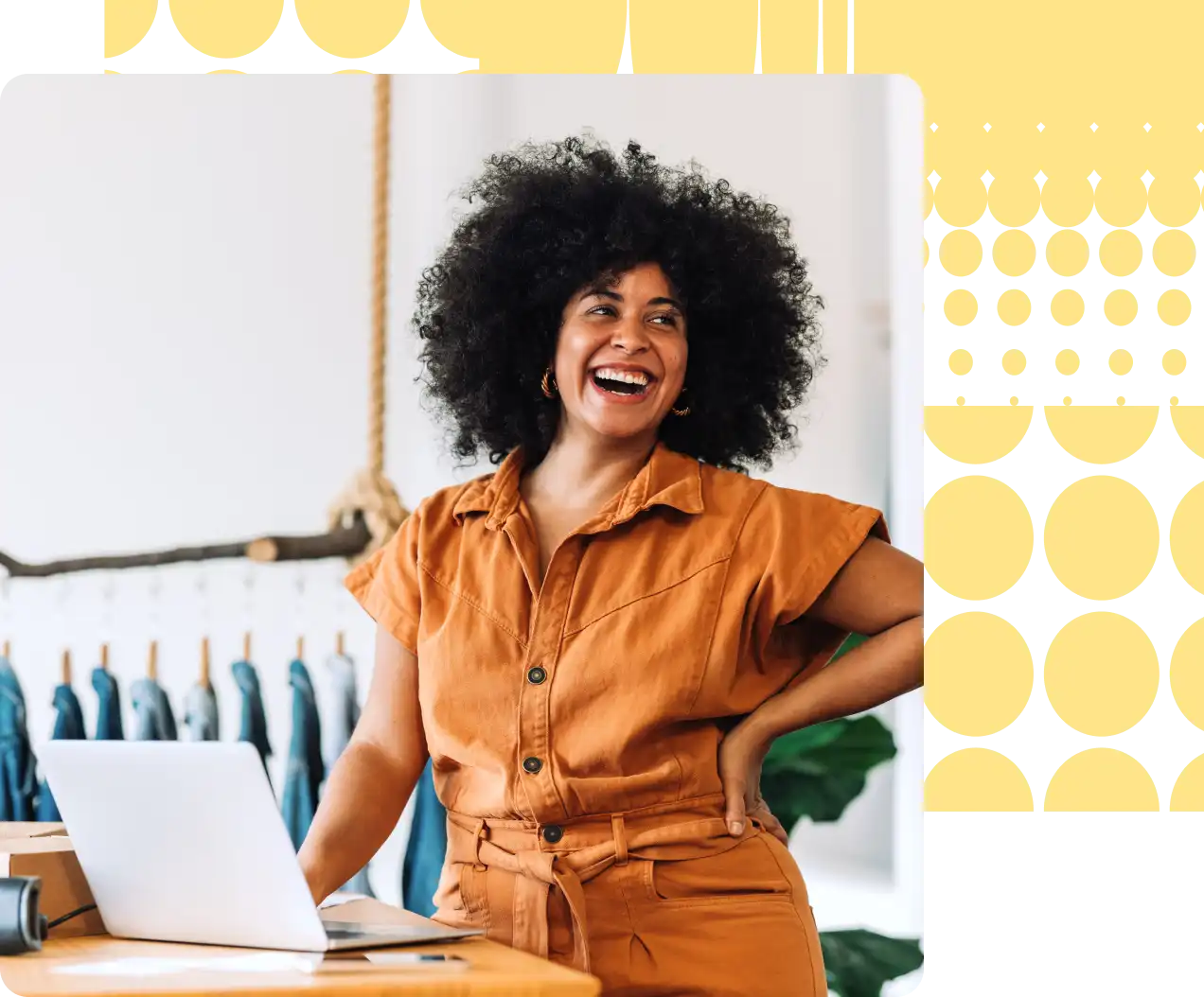 Woman stands behind a desk with a laptop on it laughing at something out of the frame